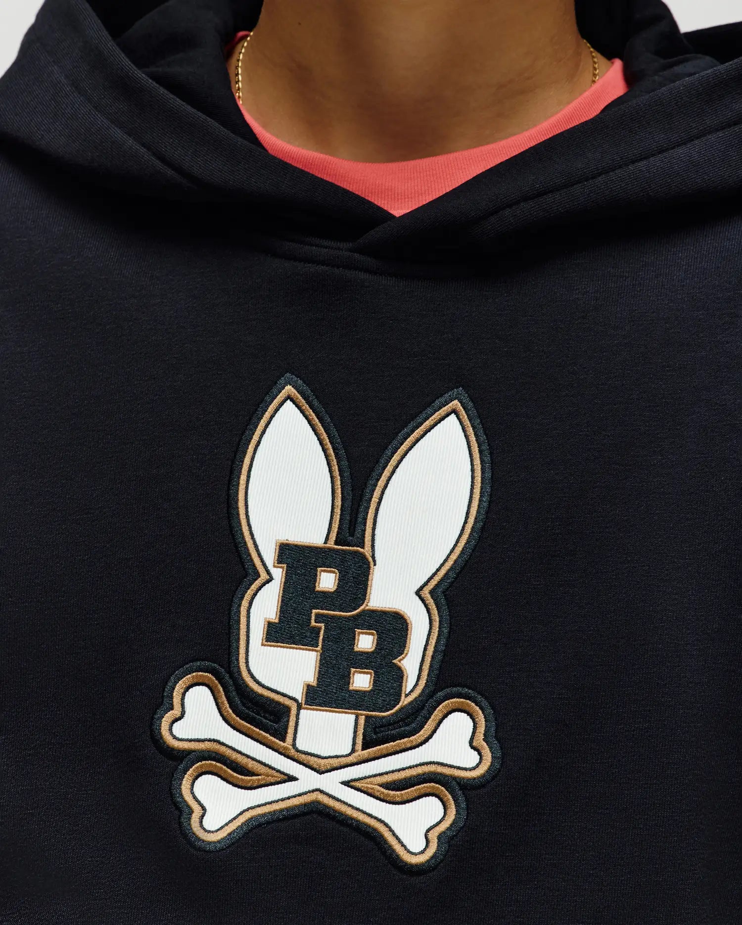 A person wears the Psycho Bunny KIDS WADE CORD PATCH HOODIE (KT0500031), made from a premium cotton blend with an embroidered Bunny patch showing bunny ears, "PB," and crossed bones. A pink shirt and gold chain peek at the neckline.