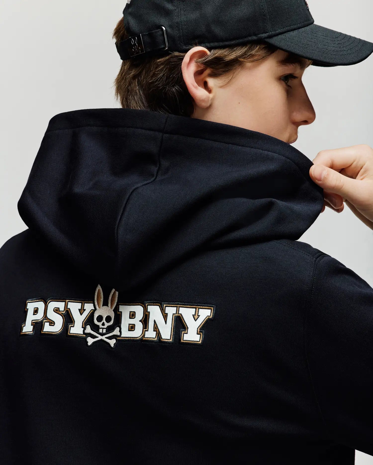 A person in the Psycho Bunny KIDS WADE CORD PATCH HOODIE (KT0500031), a black premium cotton blend, wearing a black cap and holding the hoodie near their neck. Features “PSYBNY” and bunny skull and crossbones graphic on the back.