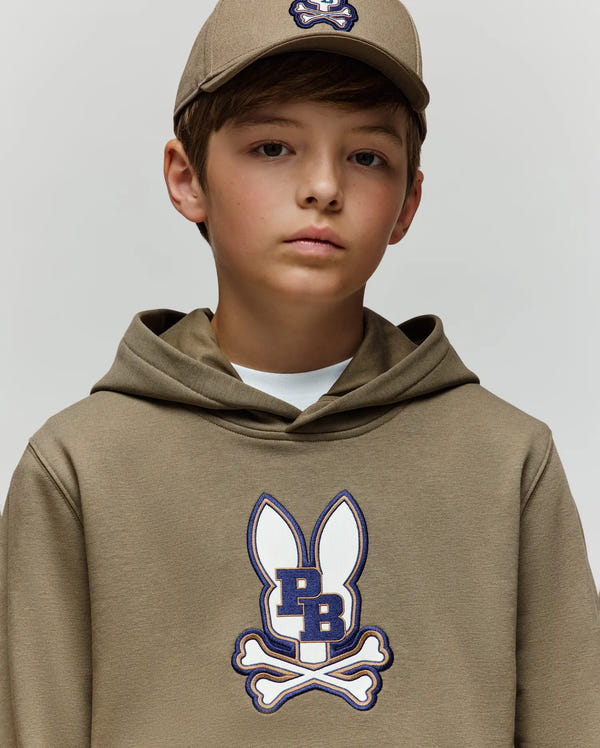A young boy wears the Psycho Bunny KIDS WADE CORD PATCH HOODIE (KT0500031) and matching cap, both in tan cotton blend, featuring a cartoon bunny logo with "PB" letters and crossed bones, standing against a plain light background.