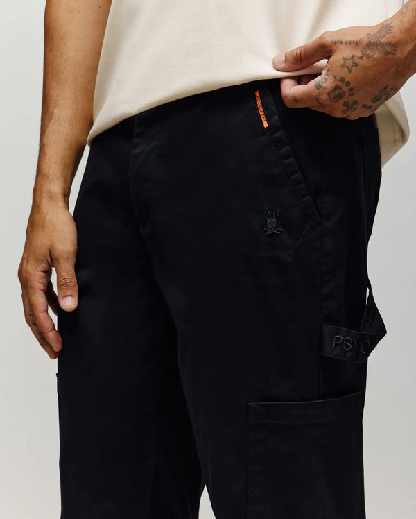 A person wearing black Psycho Bunny MENS RUSSELL CARPENTER PANT (MB0600005) with cargo pockets and a small embroidered bunny logo stands with one hand in their pocket, displaying visible hand tattoos.