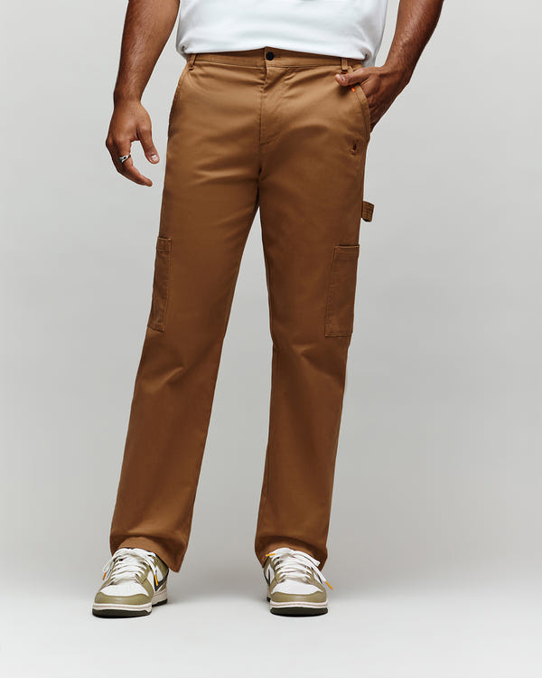 A person in Psycho Bunny MENS RUSSELL CARPENTER PANT (MB0600005) with hammer loop and side pockets, white shirt, and white sneakers with gold accents stands against a light gray background. The pants offer a regular fit for comfort and style.