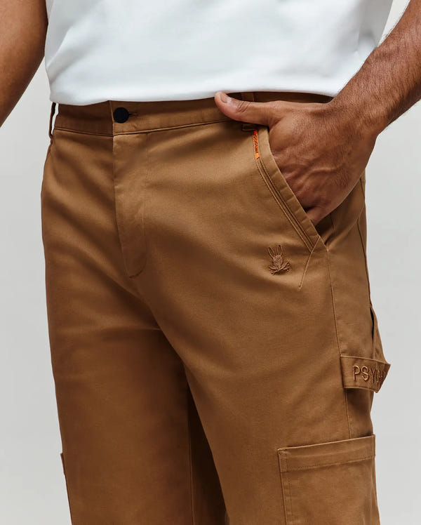A person wears tan Psycho Bunny MENS RUSSELL CARPENTER PANT (MB0600005) in a regular fit, with a white shirt tucked in. The pants feature an embroidered logo, "PSY" label on the pocket, and a functional hammer loop.