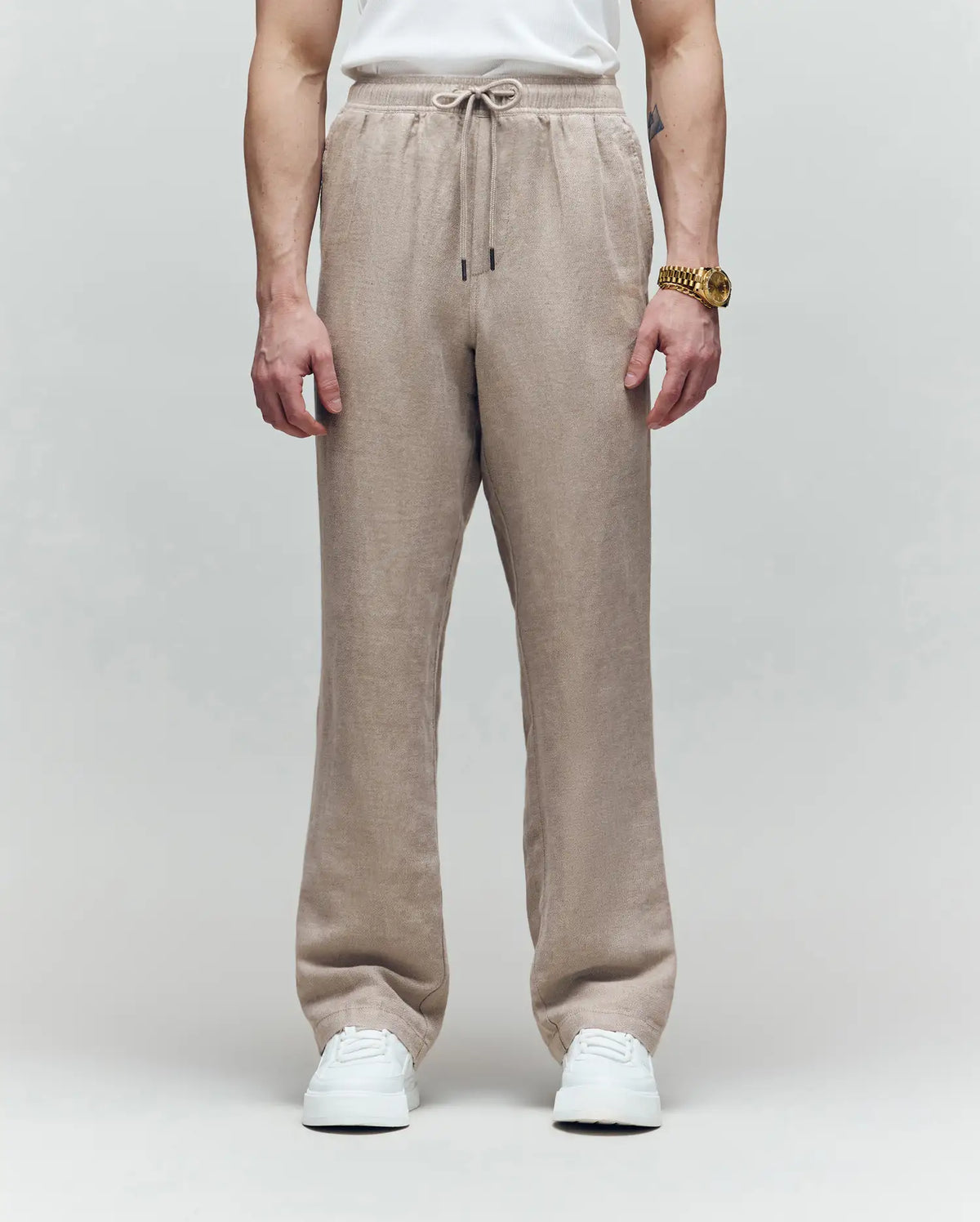 A person wearing Psycho Bunny's MENS FARRIS WIDE LEG LINEN PANT (MB0600007) in light brown, paired with a white T-shirt, white sneakers, and a gold watch stands against a gray background; only their body from the shoulders down is visible.