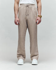 A person wearing Psycho Bunny's MENS FARRIS WIDE LEG LINEN PANT (MB0600007) in light brown, paired with a white T-shirt, white sneakers, and a gold watch stands against a gray background; only their body from the shoulders down is visible.