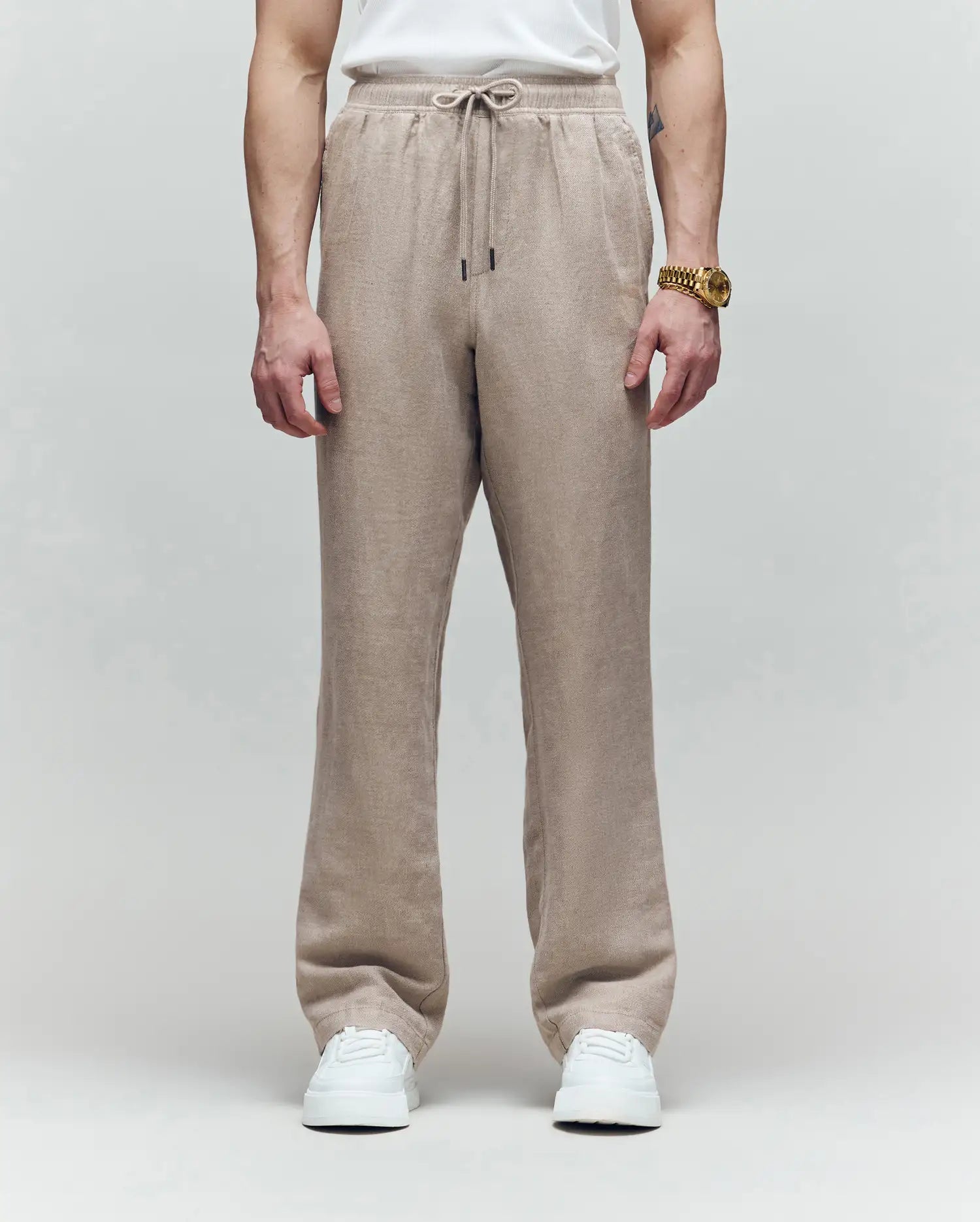 A person wearing Psycho Bunny's MENS FARRIS WIDE LEG LINEN PANT (MB0600007) in light brown, paired with a white T-shirt, white sneakers, and a gold watch stands against a gray background; only their body from the shoulders down is visible.