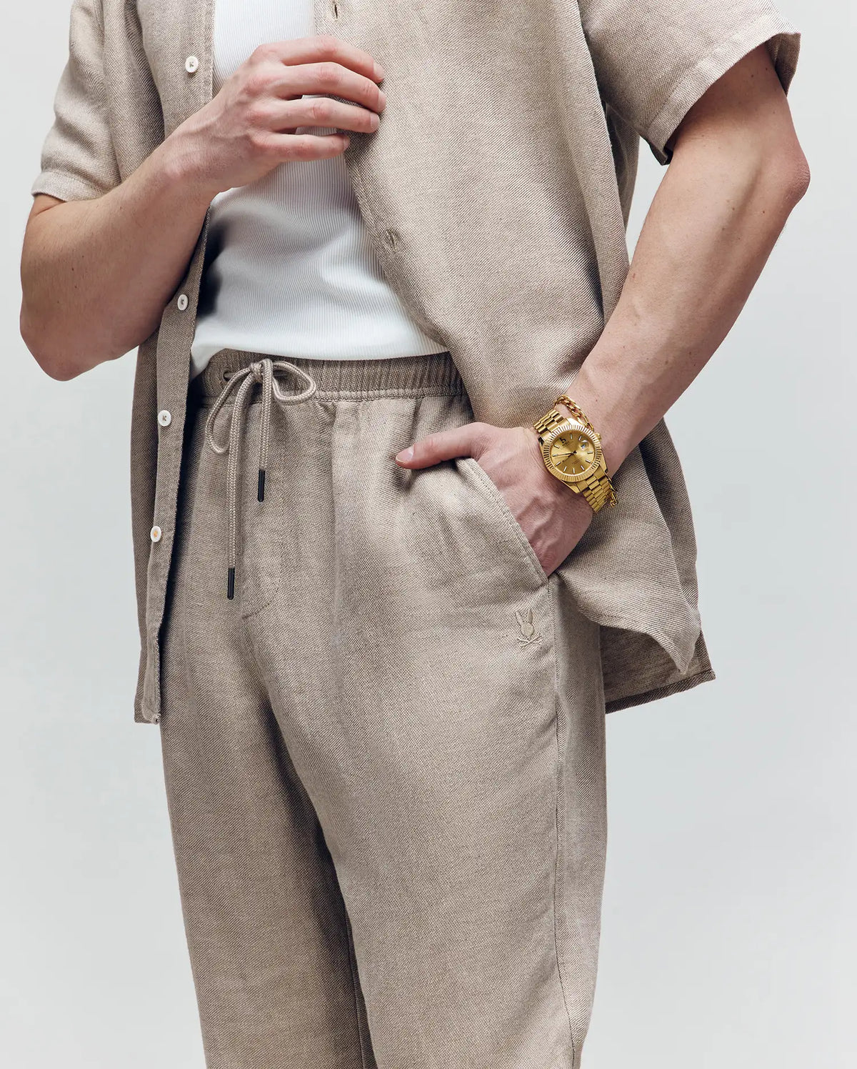 A person wears Psycho Bunny's MENS FARRIS WIDE LEG LINEN PANT in beige with a matching linen-blend shirt over a white tank top, accessorized with a large gold wristwatch; only the torso and arms are visible against a plain background.