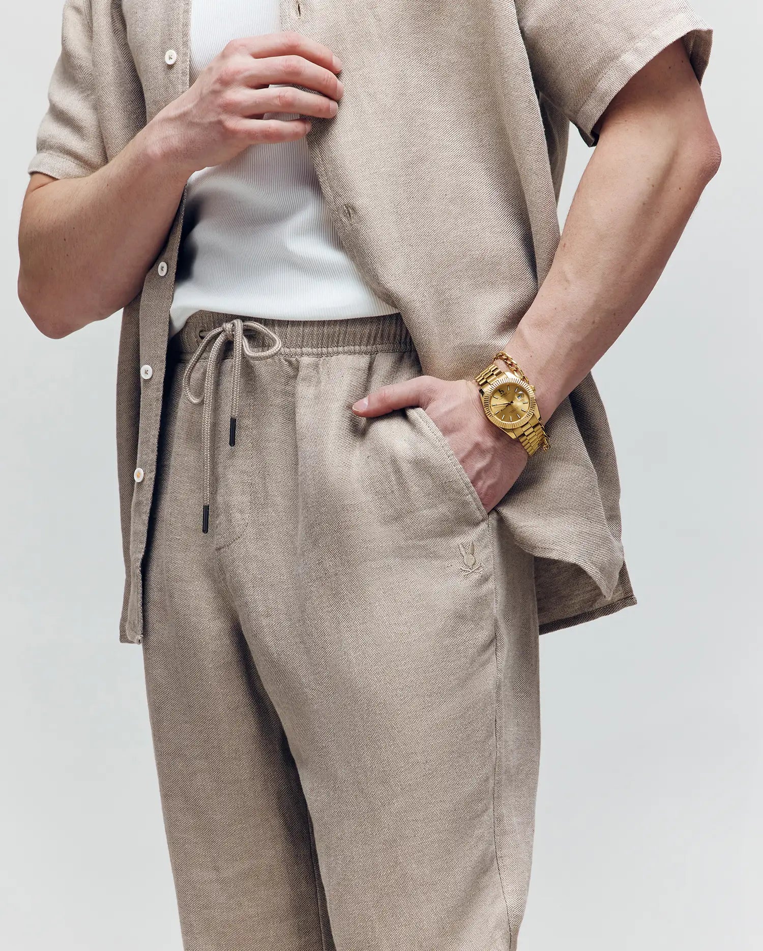 A person wears Psycho Bunny's MENS FARRIS WIDE LEG LINEN PANT in beige with a matching linen-blend shirt over a white tank top, accessorized with a large gold wristwatch; only the torso and arms are visible against a plain background.