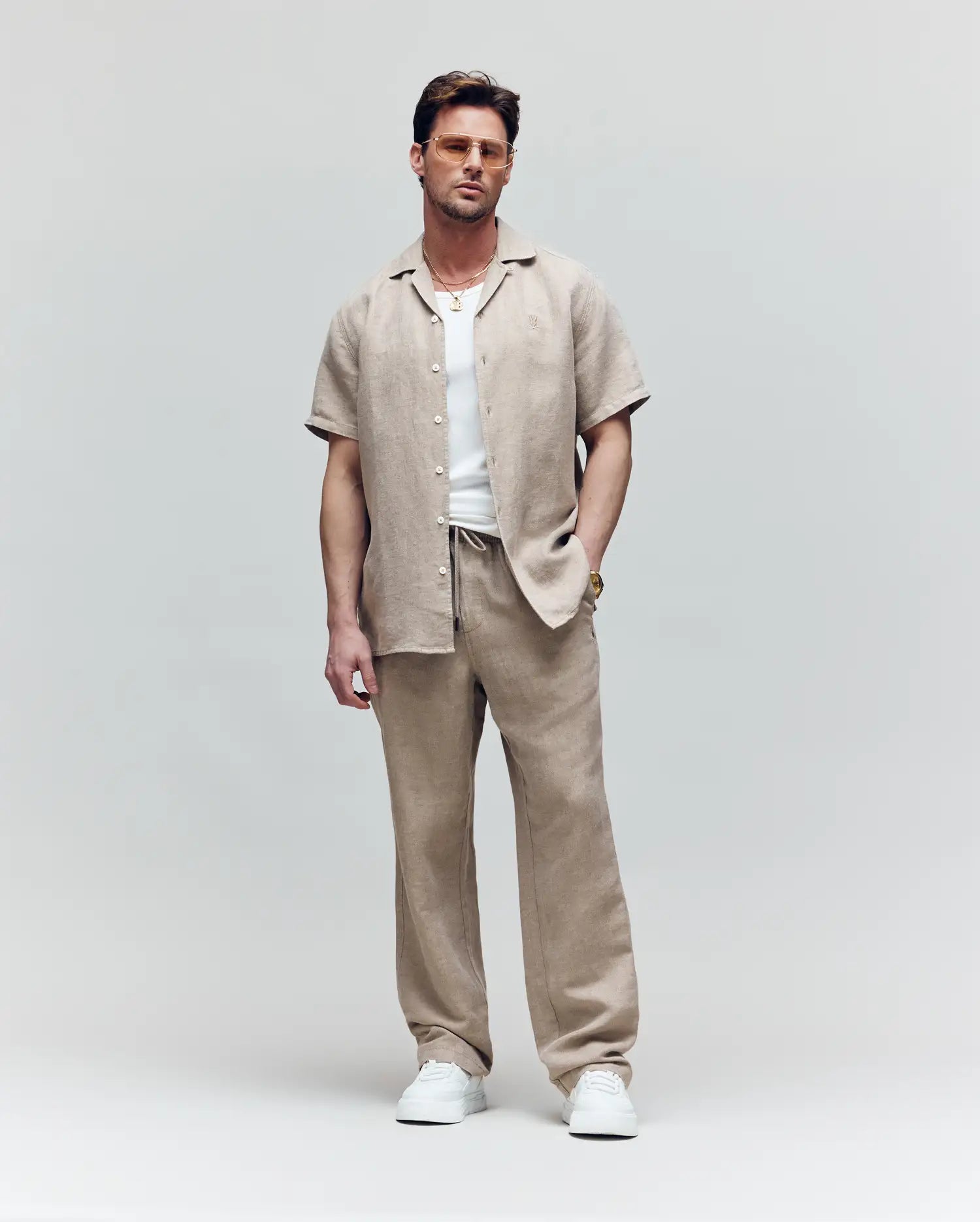 A man stands against a plain background wearing Psycho Bunny's MENS FARRIS WIDE LEG LINEN PANT (MB0600007), paired with a short-sleeve button-up shirt, white T-shirt, sneakers, sunglasses, a gold necklace, and ring, one hand in his pocket.