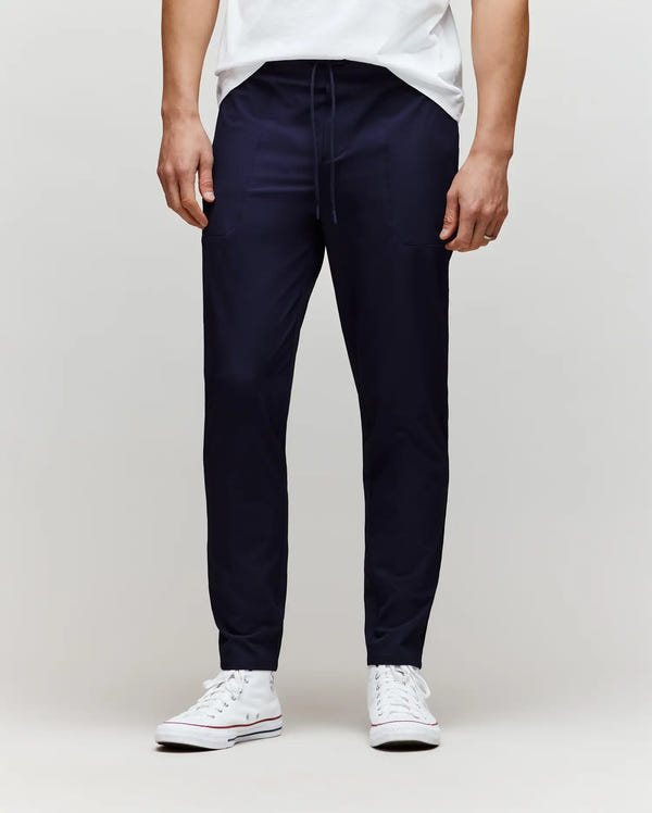 A person wears Psycho Bunny’s MENS BEN TECH JOGGER PULL ON (MB0600013) in navy, paired with a white T-shirt and white sneakers, standing against a plain light gray background. The photo is cropped from the chest down.