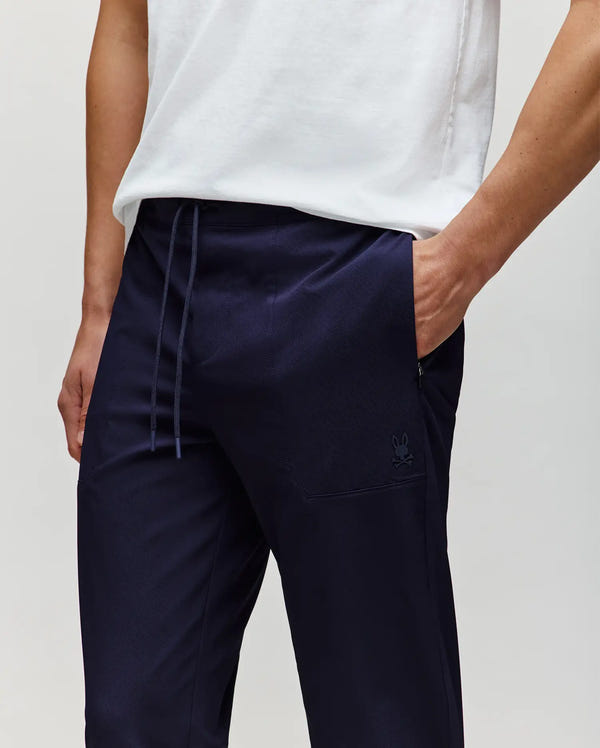 A person models Psycho Bunny’s MENS BEN TECH JOGGER PULL ON - MB0600013 in dark navy polyester spandex, shown from shoulders to mid-thigh with one hand in their pocket against a plain background.
