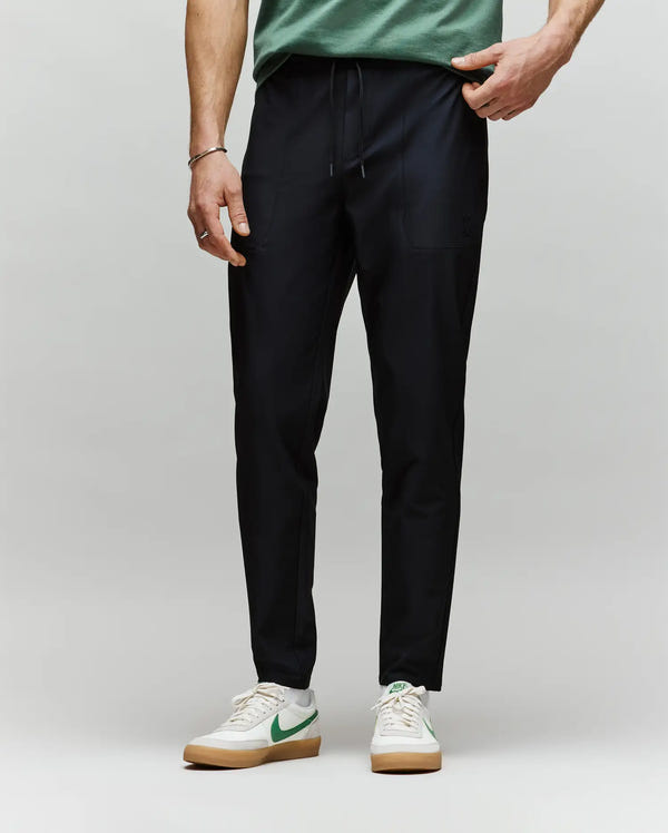 A person, visible from the waist down, stands against a plain background wearing Psycho Bunny MENS BEN TECH JOGGER PULL ON - MB0600013 in black, paired with a green shirt and white sneakers featuring green Nike swooshes.