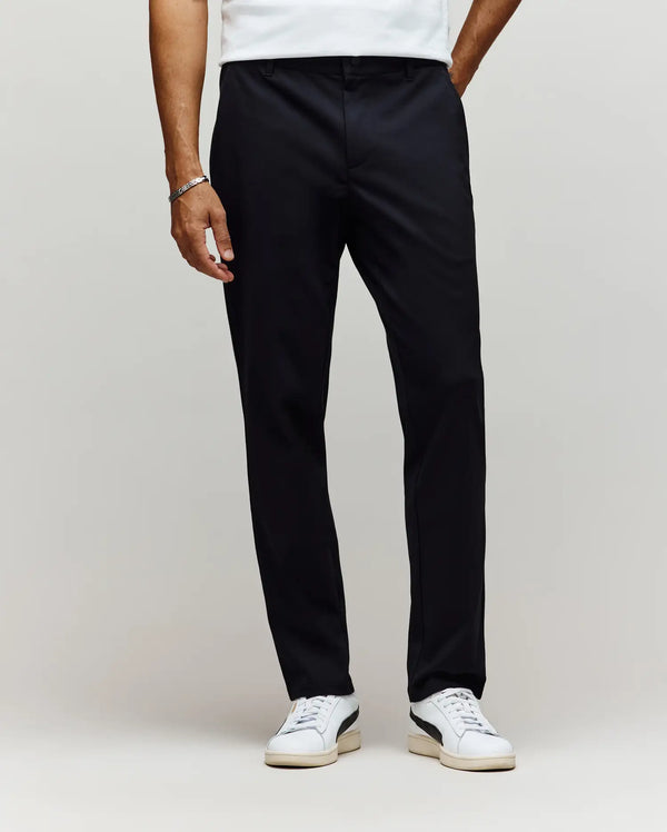 A person wears Psycho Bunny MENS EDWARD PANT - MB0600014, a white cotton woven T-shirt, white sneakers with light soles, and a silver bracelet, standing against a plain light background.