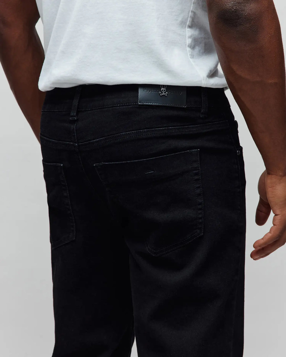 Black Mens Emerson Denim Pant, classic fit, featuring a subtle denim texture, five-pocket styling, and a small silver Psycho Bunny skull logo on a black leather patch at the rear waistband.