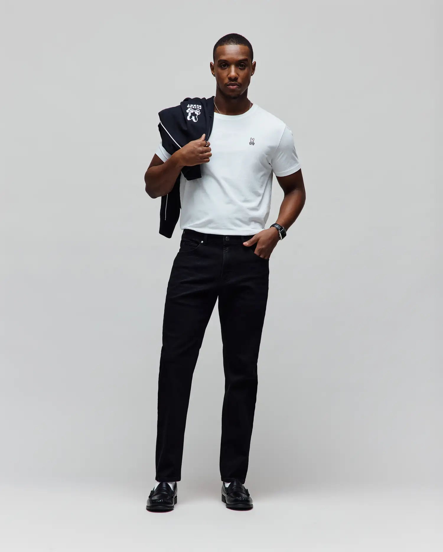 White classic fit jersey t-shirt with small embroidered Psycho Bunny logo on left chest, black track jacket featuring large white back graphic and piping, and Mens Emerson Denim Pant.