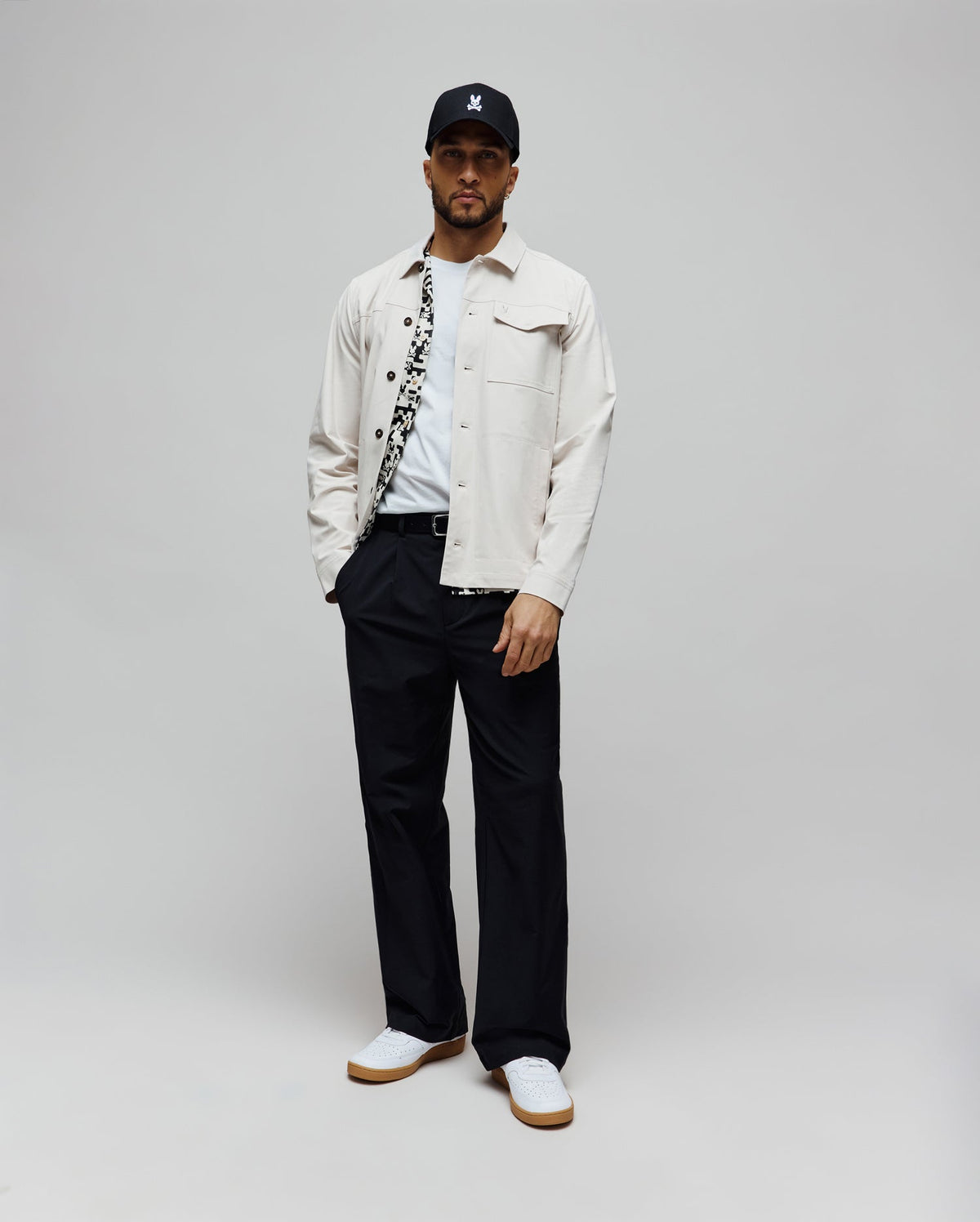 Black cap with white embroidered Psycho Bunny skull logo, light beige twill shirt jacket with patterned lining, white jersey t-shirt, and Mens Malik Wide Leg Pant in navy with pleats, paired with white gum-sole sneakers.