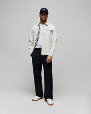Black cap with white embroidered Psycho Bunny skull logo, light beige twill shirt jacket with patterned lining, white jersey t-shirt, and Mens Malik Wide Leg Pant in navy with pleats, paired with white gum-sole sneakers.