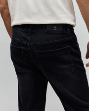 Mens Ace Knit Denim Pant in washed black, featuring a classic fit, visible back pockets, and a small metallic Psycho Bunny skull logo on a black leather-like patch at the back waistband.