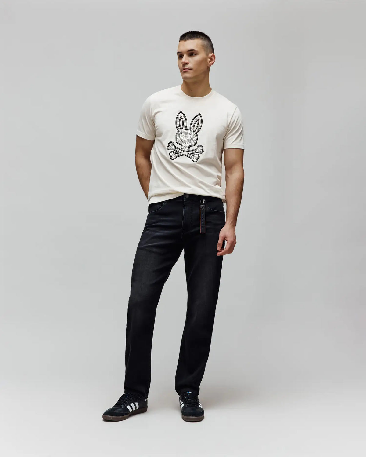 Cream jersey t-shirt with a large, intricate black Psycho Bunny skull logo on the chest, paired with dark wash Mens Ace Knit Denim Pant, relaxed fit, and a branded tag.