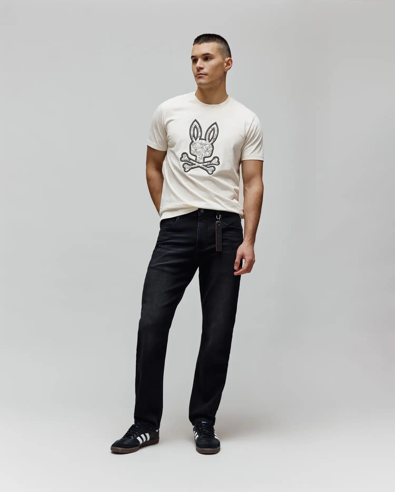 Cream jersey t-shirt with a large, intricate black Psycho Bunny skull logo on the chest, paired with dark wash Mens Ace Knit Denim Pant, relaxed fit, and a branded tag.