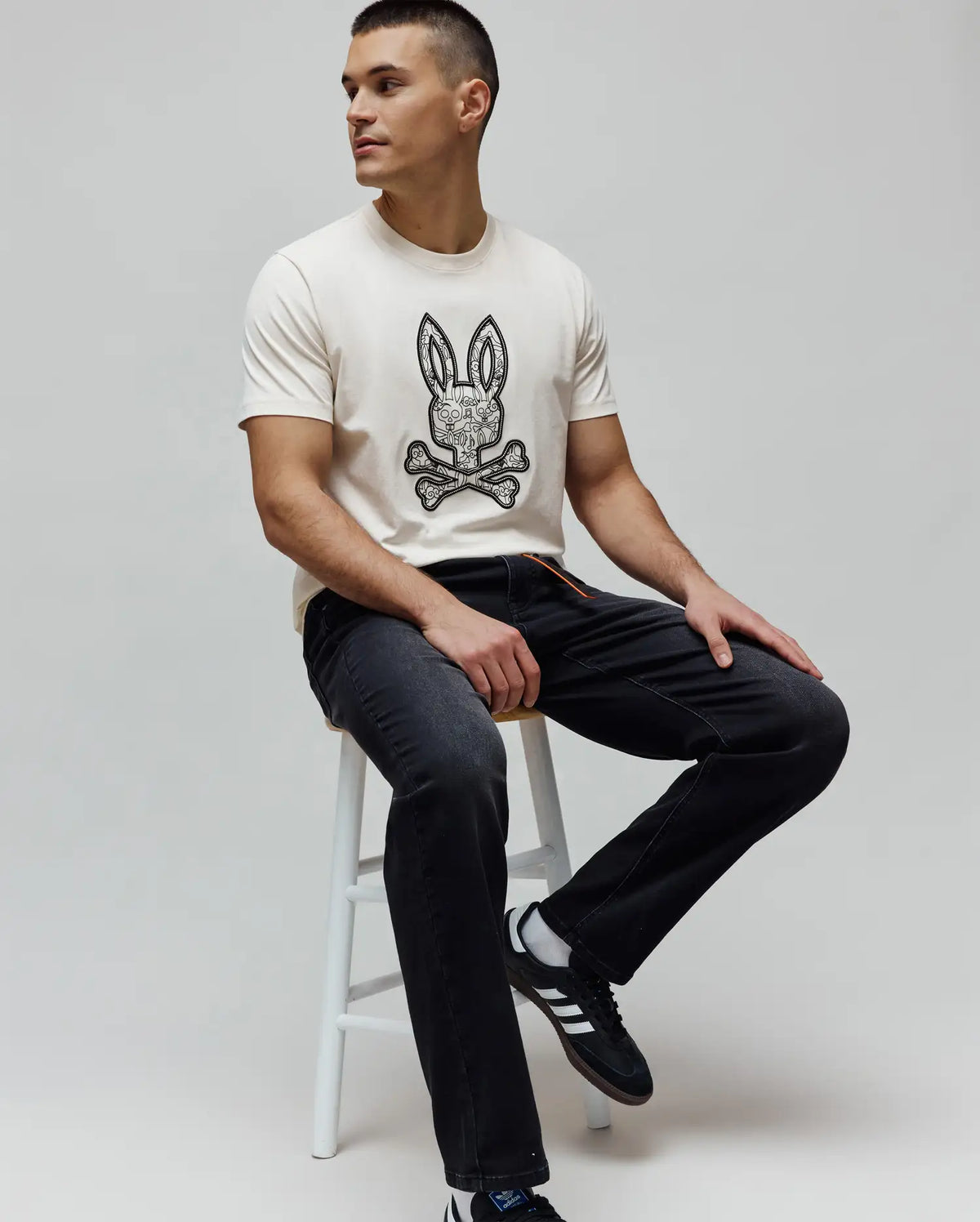 Cream jersey t-shirt with a large, intricate black and white Psycho Bunny skull and crossbones logo on the chest, paired with MENS ACE KNIT DENIM PANT in washed black with orange drawstring detail.