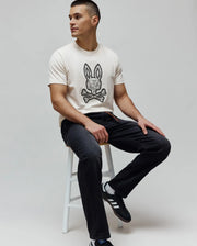 Cream jersey t-shirt with a large, intricate black and white Psycho Bunny skull and crossbones logo on the chest, paired with MENS ACE KNIT DENIM PANT in washed black with orange drawstring detail.
