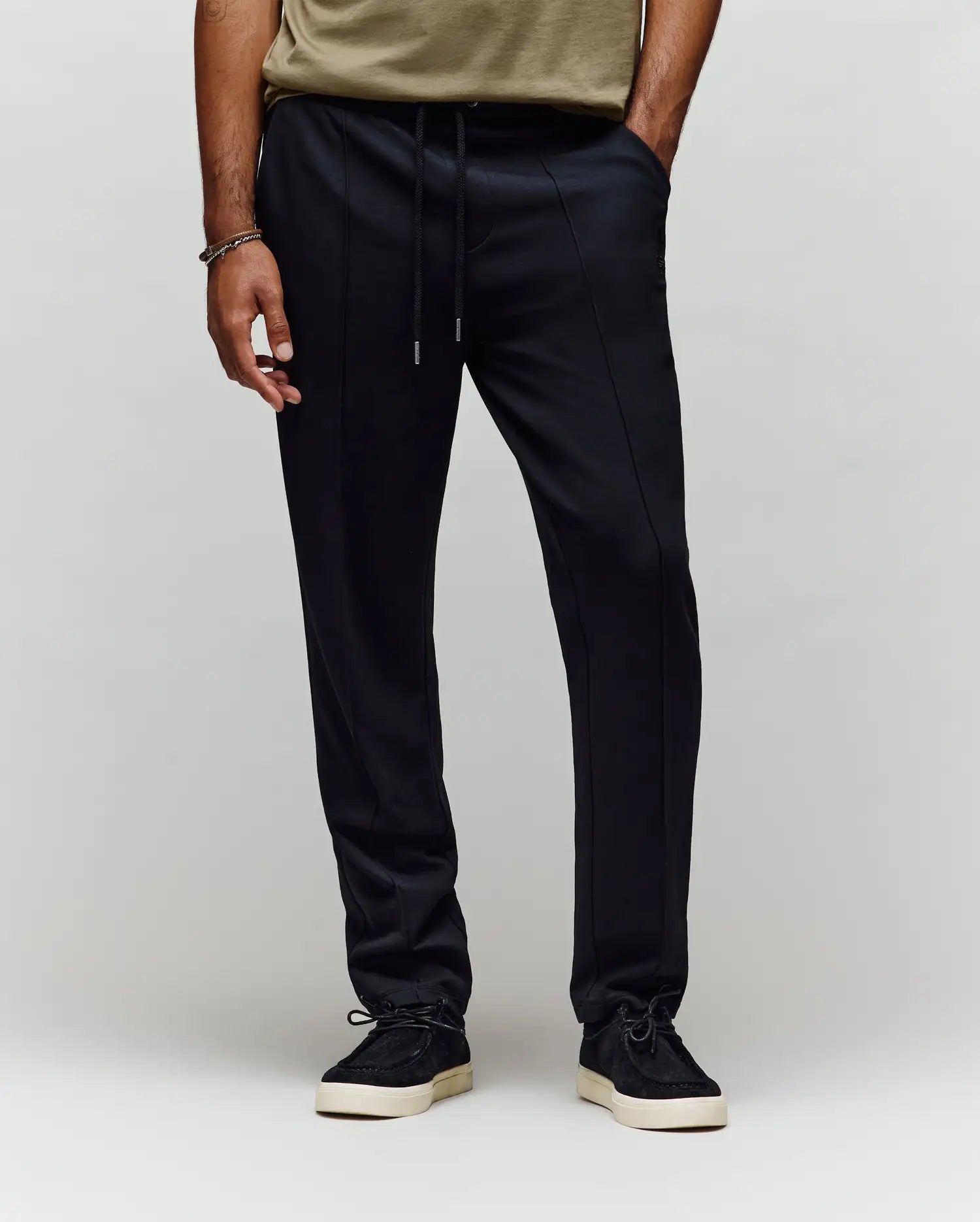 A person in a beige T-shirt, Psycho Bunny MENS ALEXIS PINTUCK JOGGER - MB0800006 black pants with seam details and bunny logo, black sneakers with white soles, and a bracelet, stands against a plain light background.