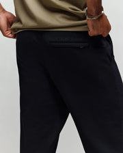 A person is seen from the waist down wearing Psycho Bunny's MENS ALEXIS PINTUCK JOGGER - MB0800006 in black, with "Psycho Bunny" embroidery above the back pocket, paired with a khaki t-shirt and one hand resting near the pocket.