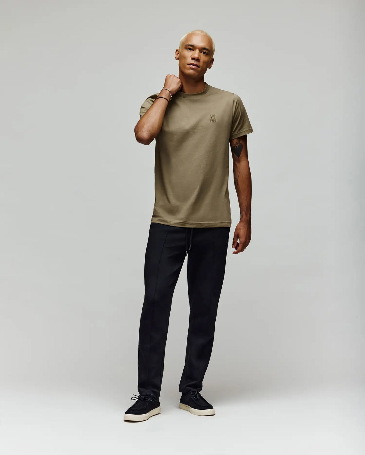 A person with short blond hair stands against a plain light background, wearing a khaki t-shirt and black Psycho Bunny MENS ALEXIS PINTUCK JOGGER (MB0800006), with one hand resting on their neck, looking relaxed.