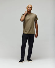 A person with short blond hair stands against a plain light background, wearing a khaki t-shirt and black Psycho Bunny MENS ALEXIS PINTUCK JOGGER (MB0800006), with one hand resting on their neck, looking relaxed.