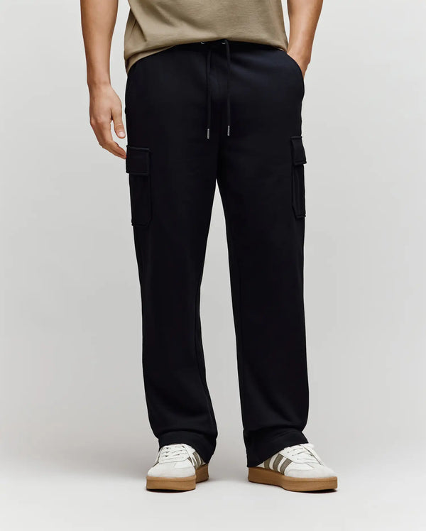 Wearing Psycho Bunny MENS NASH CARGO SWEATPANT - MB0800010, black with roomy cargo pockets and sweatpant comfort, paired with white/beige sneakers and a beige shirt. The background is light; only the lower body is fully visible.