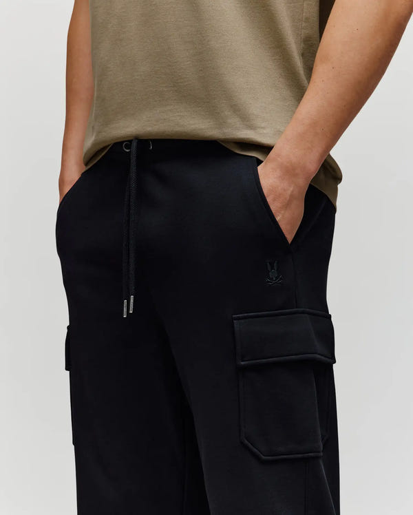 A person wears a khaki t-shirt and Psycho Bunny MENS NASH CARGO SWEATPANT - MB0800010, hands in pockets, shown from shoulders to mid-thigh against a plain background, combining classic style with the comfort of sweatpants.