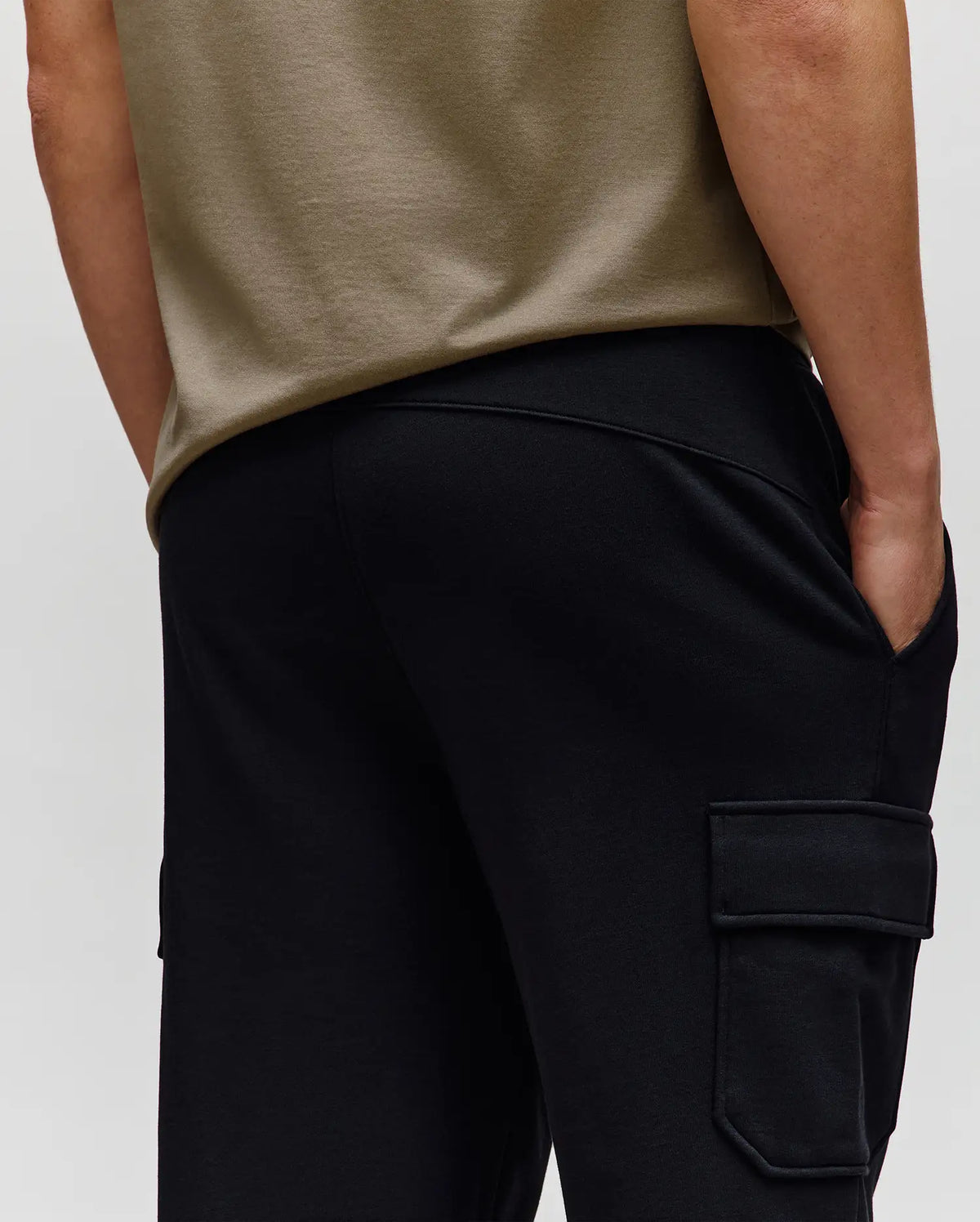 A person is shown from the back wearing a beige t-shirt and Psycho Bunny MENS NASH CARGO SWEATPANT - MB0800010, one hand in a pocket, highlighting both its utility cargo pockets and comfortable sweatpant style.