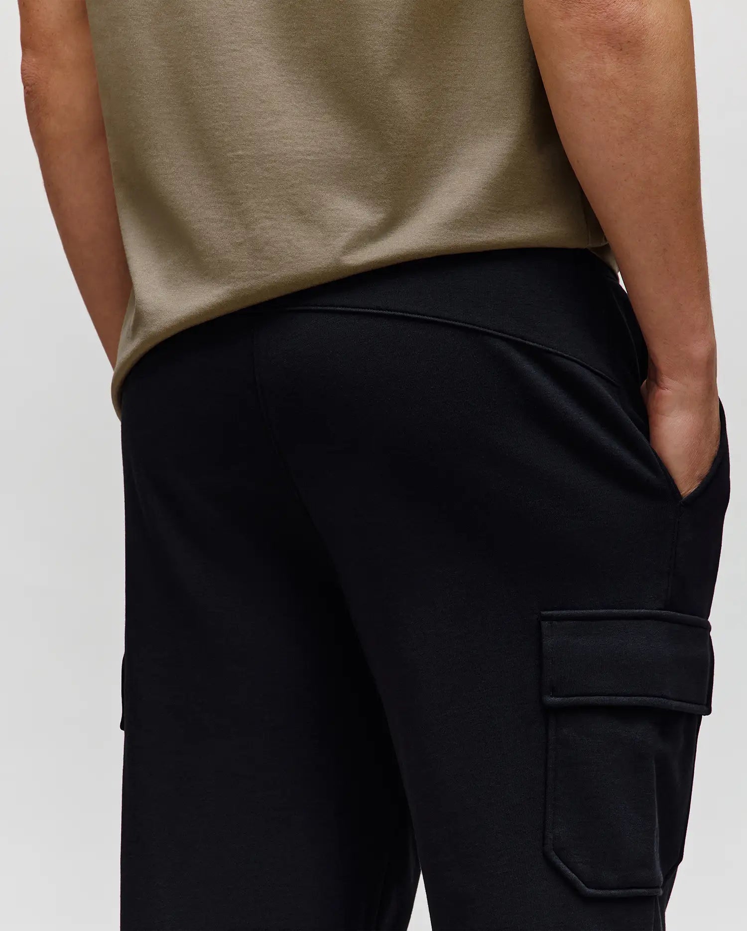 A person is shown from the back wearing a beige t-shirt and Psycho Bunny MENS NASH CARGO SWEATPANT - MB0800010, one hand in a pocket, highlighting both its utility cargo pockets and comfortable sweatpant style.