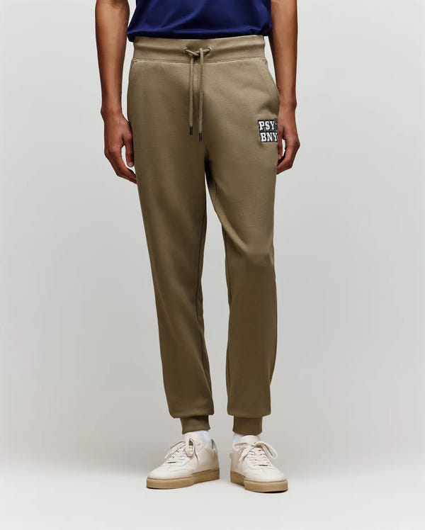 A person wears Psycho Bunny MENS WADE CORD PATCH SWEATPANT - MB0800014 in olive green, featuring "PSY BNK" on the left thigh, rib knit cuffs, and an athletic fit, paired with a navy shirt and white sneakers against a plain background.