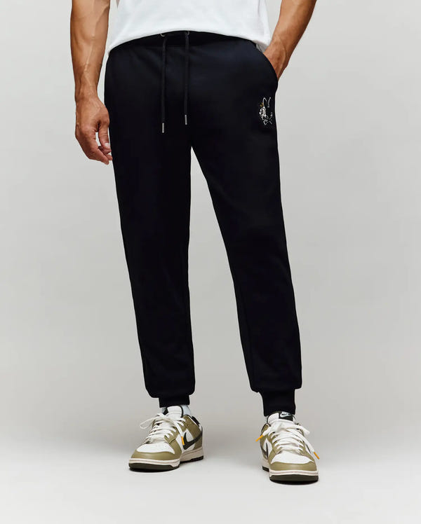 A person stands against a plain light gray background wearing Psycho Bunny MENS AUGUSTE JOGGER - MB0800015 in black with a small chenille patch on the left thigh, a white T-shirt, and beige-and-white sneakers.