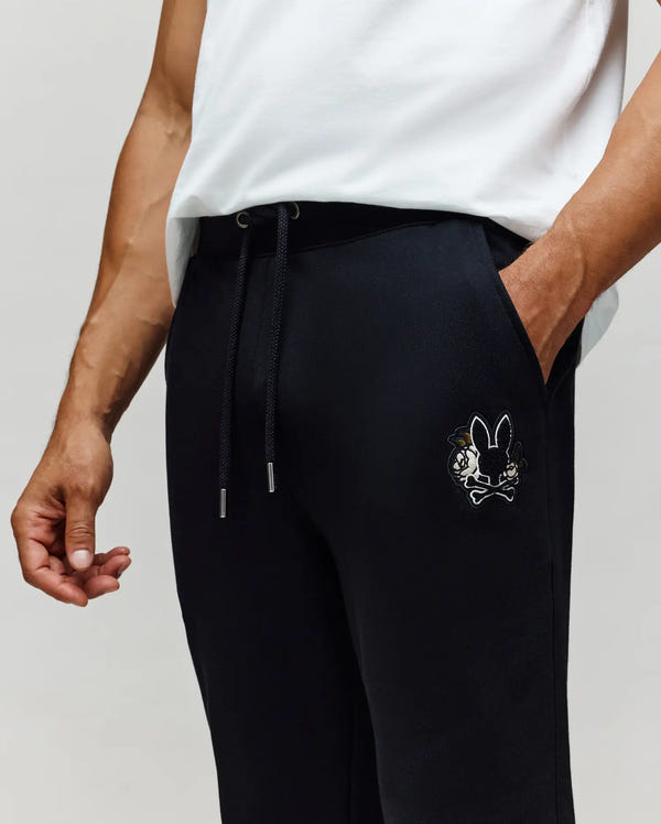 A person is shown from the waist down, wearing Psycho Bunny MENS AUGUSTE JOGGER - MB0800015 in black 100% cotton, featuring a drawstring waist and an embroidered cartoon bunny with crossbones logo on the pocket. Only one hand is visible.
