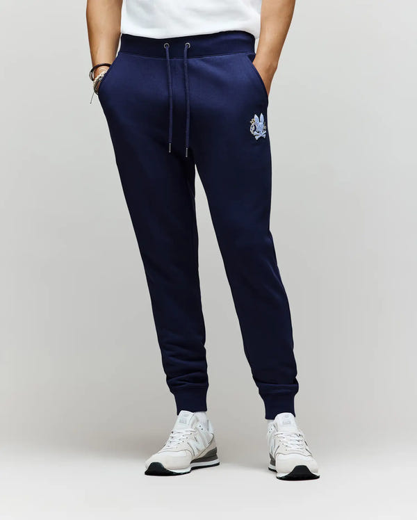 A person wears Psycho Bunny MENS AUGUSTE JOGGER - MB0800015 in navy blue, made from 100% cotton with a small chenille patch on the left thigh, paired with white sneakers and t-shirt; hands in pockets, against a plain light background.