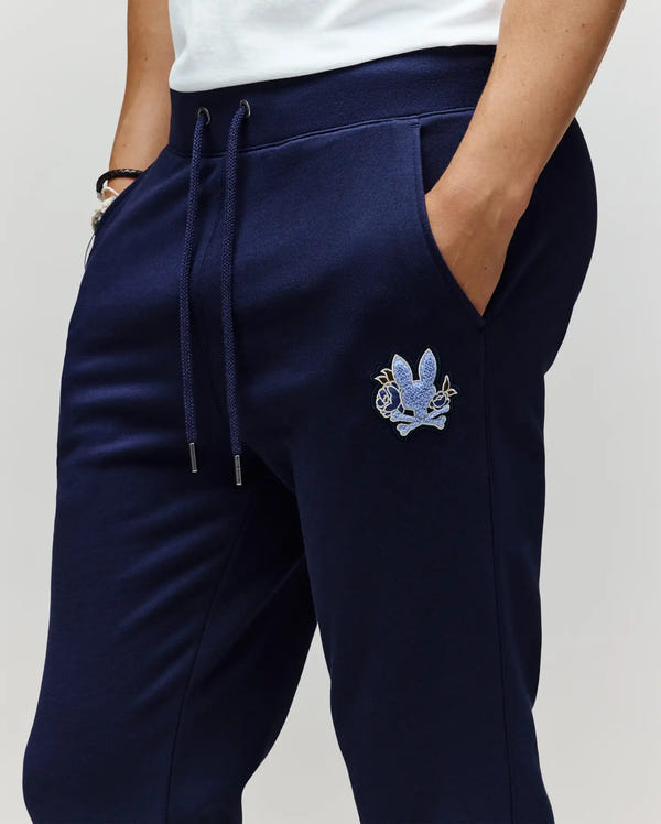 A person wears Psycho Bunny's MENS AUGUSTE JOGGER - MB0800015 in navy blue, featuring a blue chenille patch with a stylized graphic on the upper thigh, one hand in the pocket, paired with a white shirt. Made from 100% cotton.