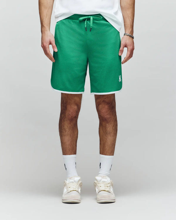 A person wears Psycho Bunny's MENS NICO MESH SHORT - MB0900004 in green with white trim and a small white logo on the left leg, styled with white socks and sneakers, standing against a plain light gray background.