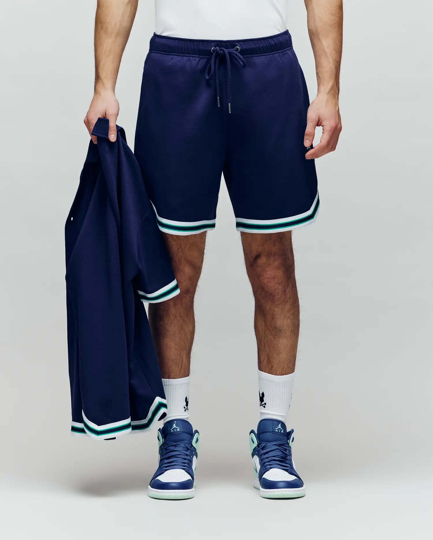 A person wears Psycho Bunny’s MENS BASIL TAPED SHORT (MB0900005)—a cotton-modal blend with white and teal trim—paired with white socks, teal/white sneakers, and a navy jacket, radiating classic confidence against a plain background.