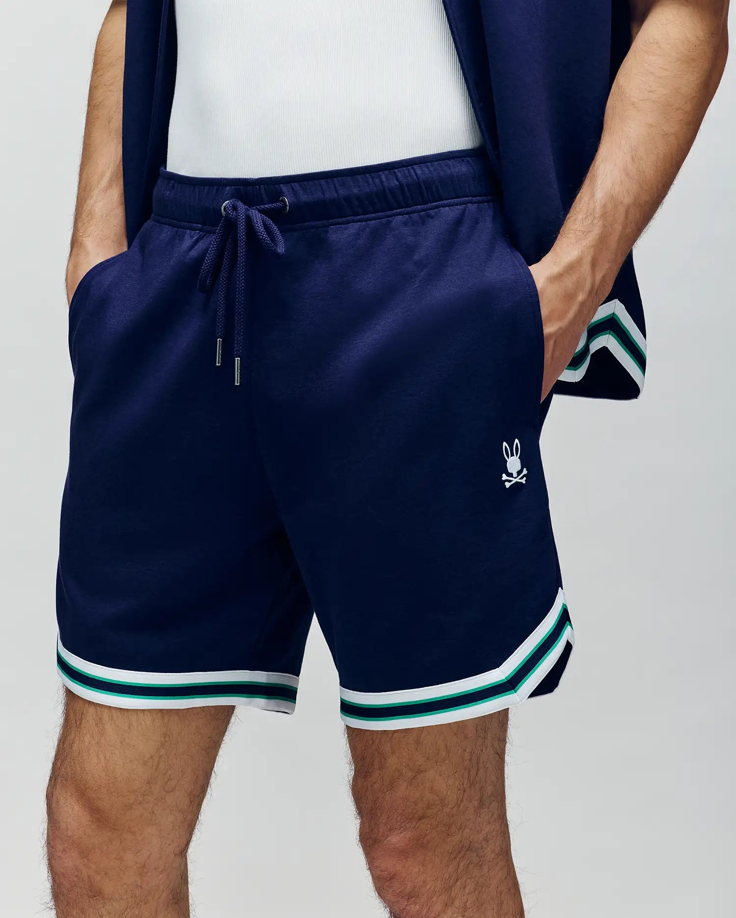 A person wears Psycho Bunny MENS BASIL TAPED SHORT (MB0900005) in navy with white and green striped trim, a small white bunny logo, a matching navy jacket, and a white tank top. Their hands are in the shorts' pockets.