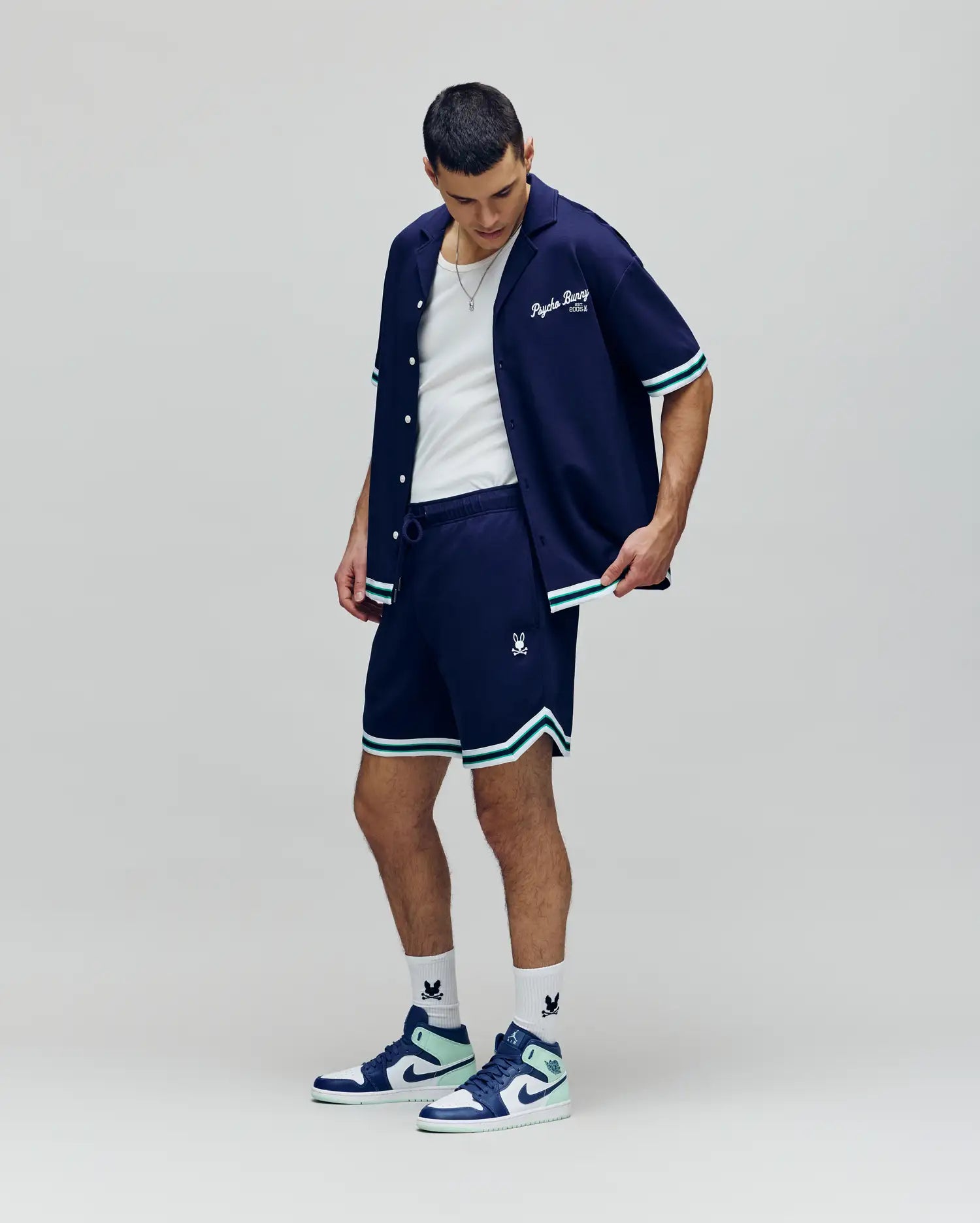 A man stands confidently in a Psycho Bunny navy button-up, MENS BASIL TAPED SHORT (MB0900005) with white and teal trim, a white tank top, logo socks, and blue and white sneakers against a plain light background.