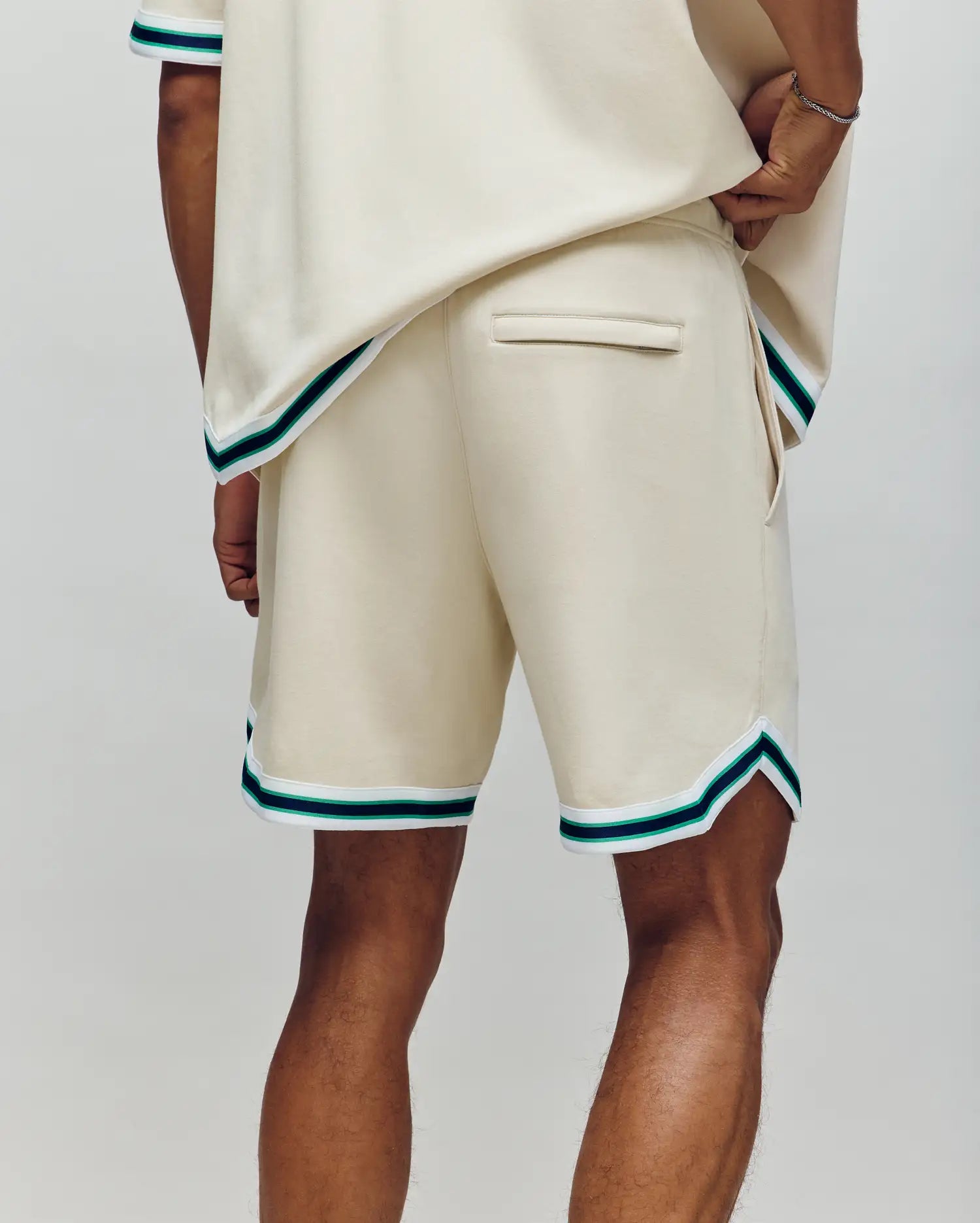 Wearing Psycho Bunny’s MENS BASIL TAPED SHORT (MB0900005)—a cream cotton-modal blend with green and black striped trim and back pocket—a person stands confidently, hand on hip, facing away from the camera.