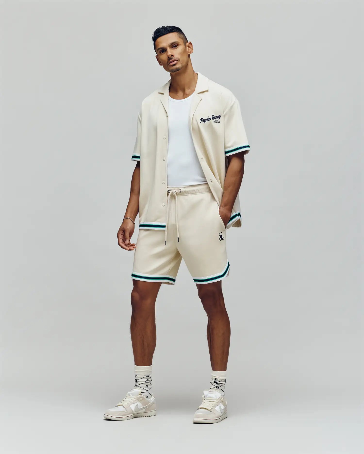 A man stands confidently against a plain background in Psycho Bunny MENS BASIL TAPED SHORT - MB0900005, paired with a cream short-sleeve shirt, white tank top, white sneakers, and patterned socks.