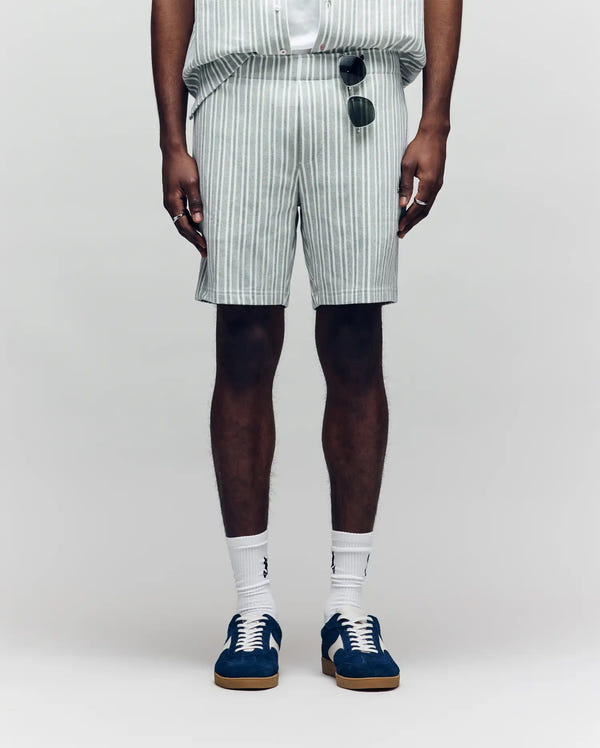 A person wears Psycho Bunny’s MENS ROBERT KNIT STRIPE SHORT - MB0900007 in light gray and white stripes, paired with a matching striped shirt, white socks, blue sneakers, and sunglasses hanging from the waistband.