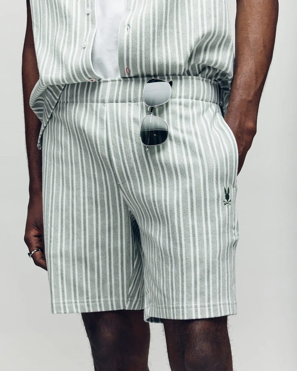 A person wearing Psycho Bunny’s MENS ROBERT KNIT STRIPE SHORT - MB0900007 in light grey and white vertical stripes, paired with a matching knit shirt; only their lower torso and upper legs are visible. Sunglasses hang from the waistband.