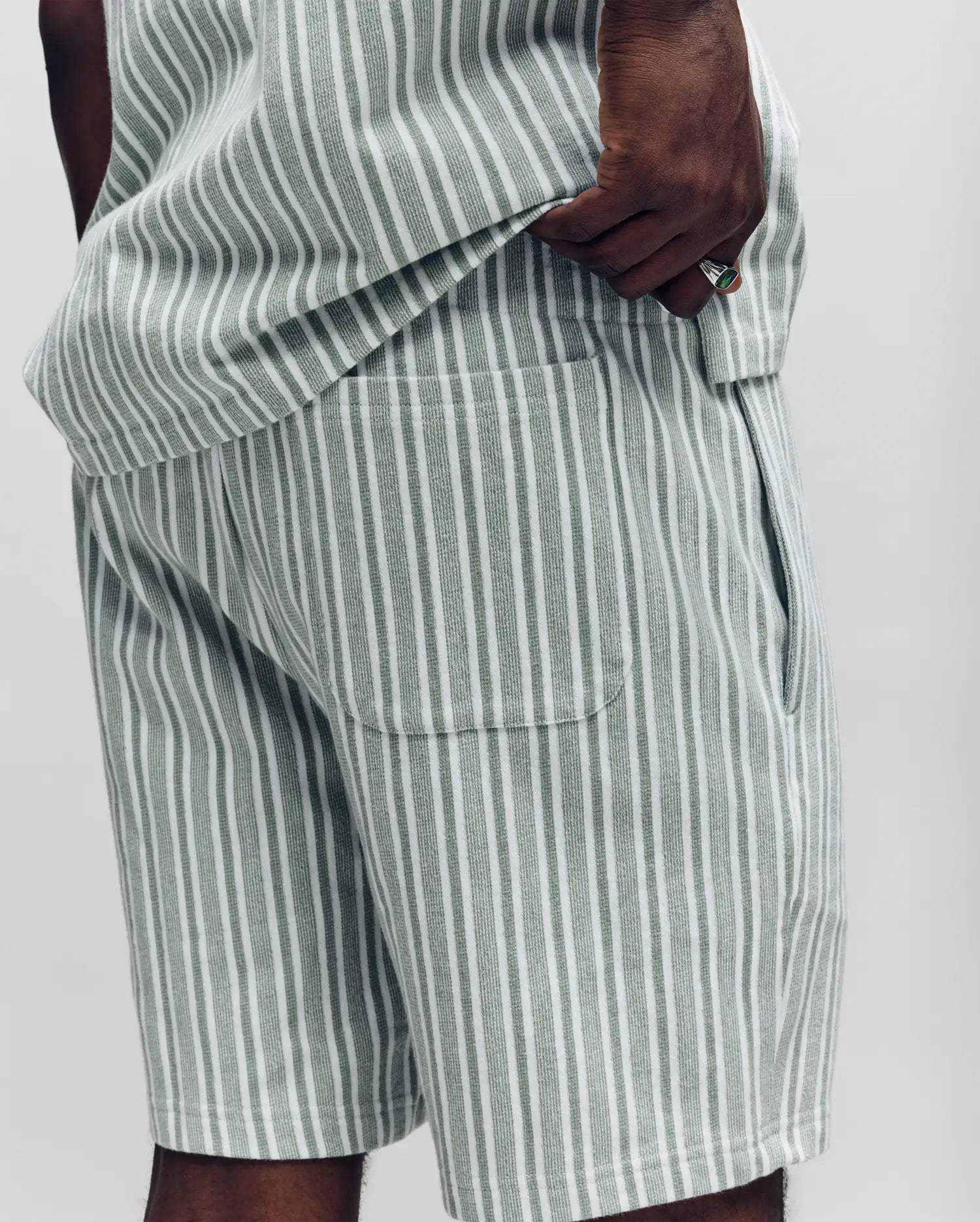 A person wears Psycho Bunny's MENS ROBERT KNIT STRIPE SHORT (MB0900007) in light gray and white stripes, paired with a matching knit cotton top. The photo, cropped to the torso and upper legs, shows one hand tucked in a pocket.