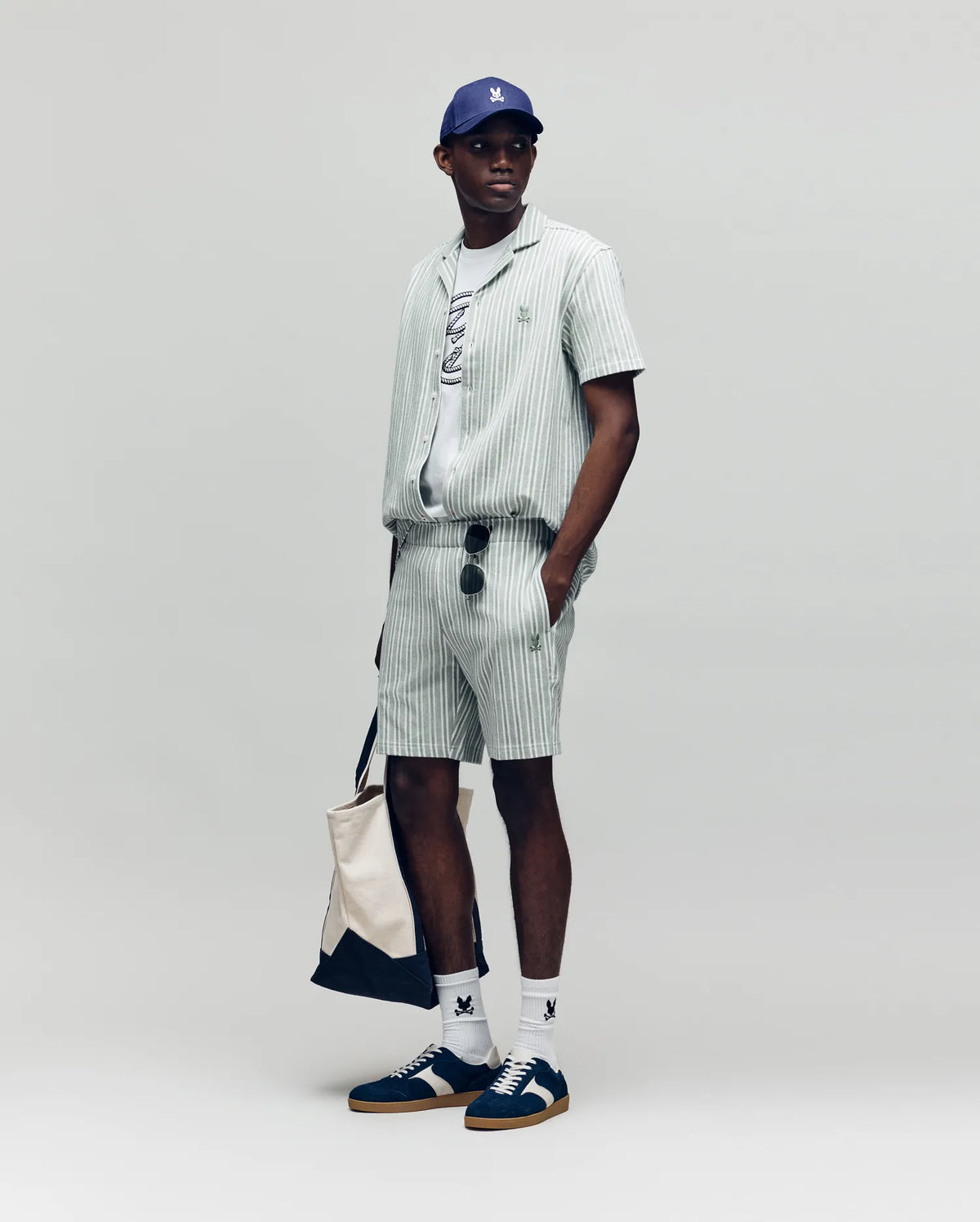 A man stands against a plain background wearing Psycho Bunny MENS ROBERT KNIT STRIPE SHORT - MB0900007 with a matching shirt, graphic tee, blue cap, white socks, dark sneakers, sunglasses, and carrying a beige tote bag.