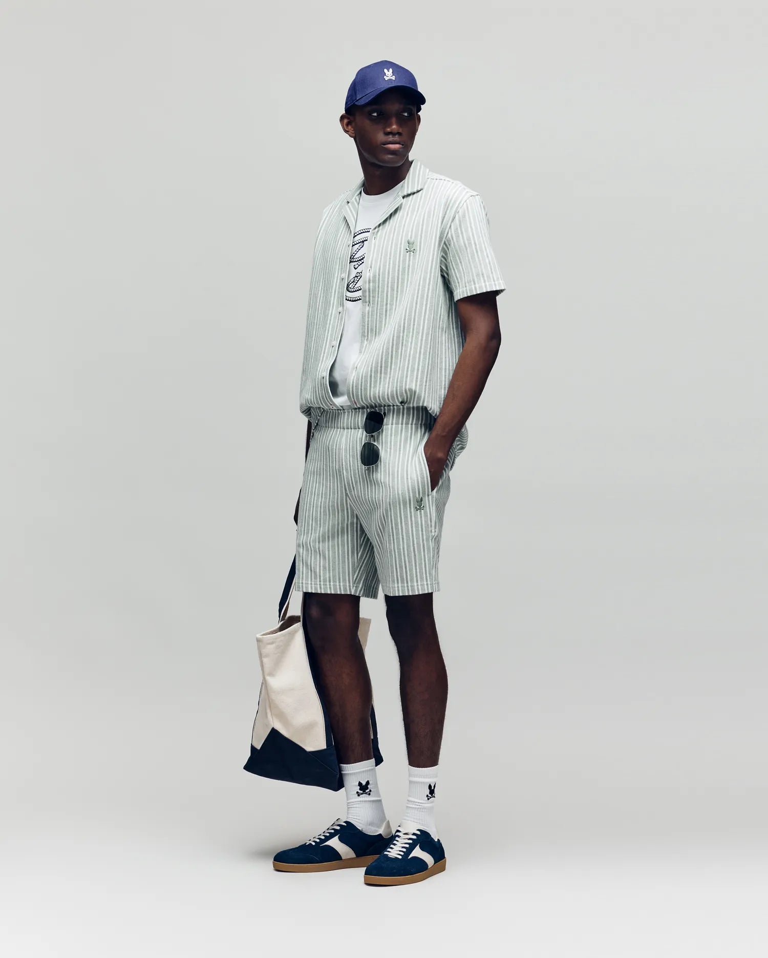 A man stands against a plain background wearing Psycho Bunny MENS ROBERT KNIT STRIPE SHORT - MB0900007 with a matching shirt, graphic tee, blue cap, white socks, dark sneakers, sunglasses, and carrying a beige tote bag.