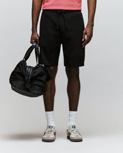 A person stands against a plain light background, wearing Psycho Bunny’s MENS RON WAFFLE CARGO SHORT (MB0900012) in black, paired with a coral waffle knit shirt, white socks, white sneakers with red accents, and holding a black duffel bag.